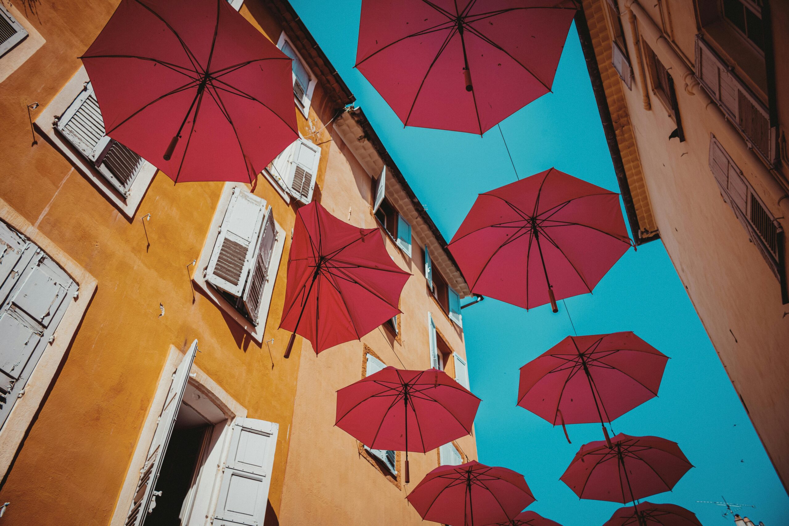 Red umbrellas suspended over a street create a vibrant contrast with a bright blue sky and architectural background.