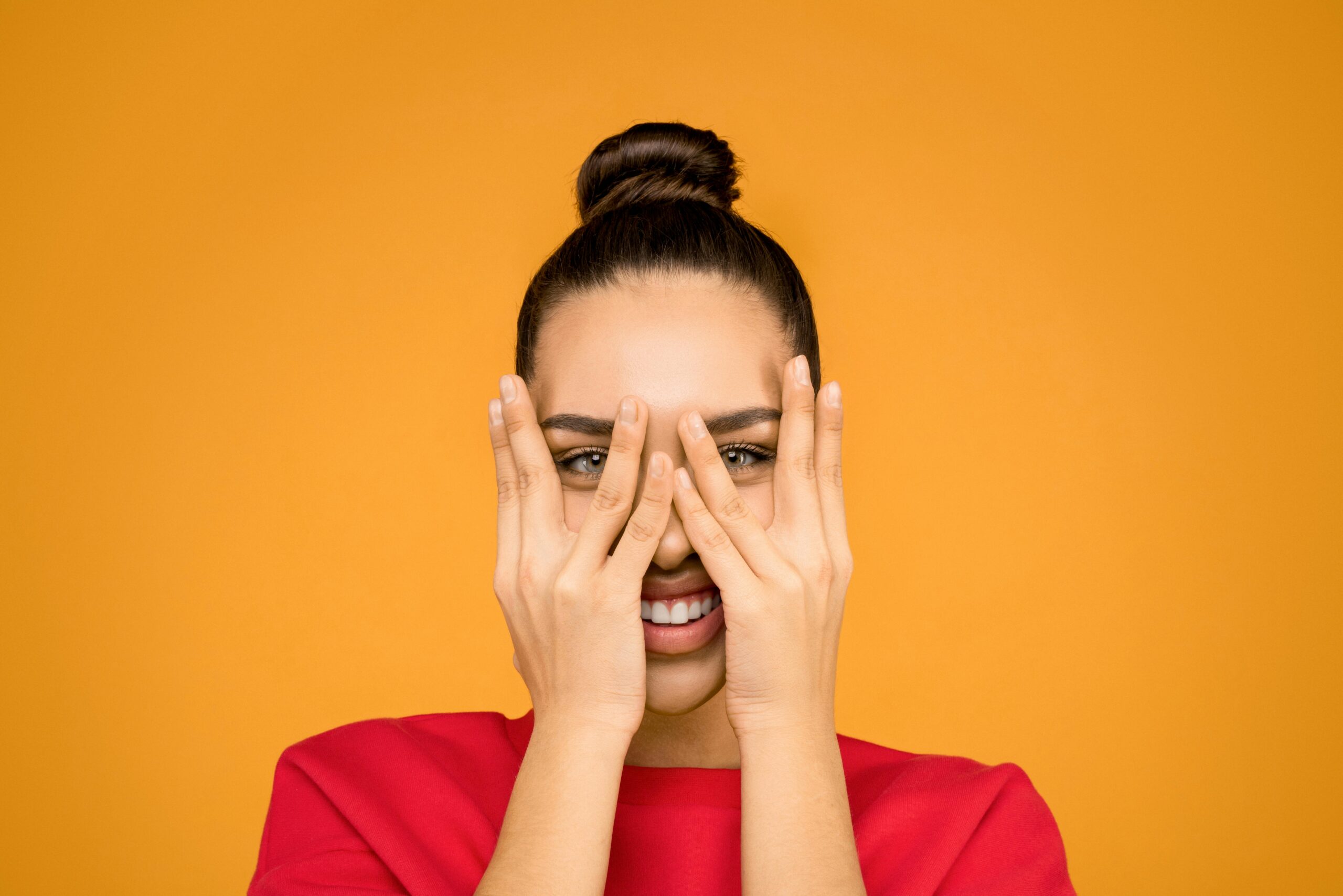A joyful woman wearing a red sweater playfully covering her face with hands against a yellow background.